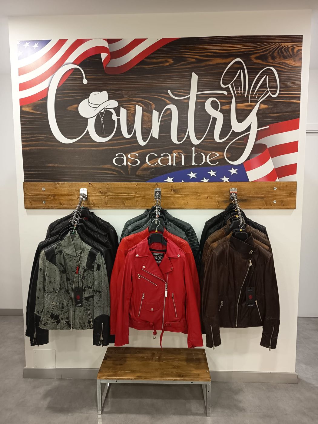 Parete interna del negozio con l&rsquo;insegna Country as can be su fondo di legno e bandiera americana, e tre giacche in pelle appese sotto